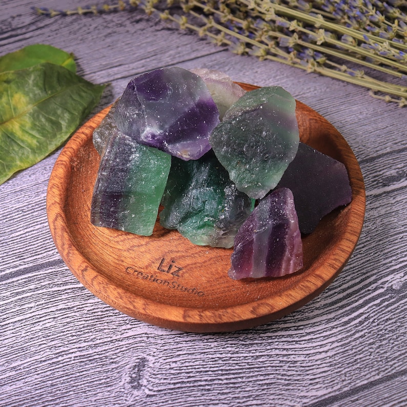 Puede incluir: Un cuenco de madera lleno de cristales de fluorita en bruto. Los cristales muestran una mezcla de tonos verdes y morados. El cuenco tiene una veta de madera natural y el texto "Liz CreationStudio" es visible.