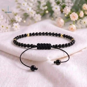 May include: A black beaded bracelet featuring small, round, polished black beads interspersed with tiny gold-colored metal spacers. The bracelet has an adjustable sliding knot closure made of black cord, with two dangling black beads at the ends.