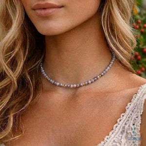 May include: A choker necklace featuring small, round, grey-toned beads with subtle swirling patterns, interspersed with tiny, polished gold-colored metallic beads. The necklace sits closely around the neck, showcasing a delicate and uniform bead arrangement.