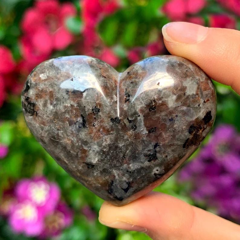 Puede incluir: Una talla de piedra en forma de coraz&oacute;n con un patr&oacute;n moteado de gris, marr&oacute;n y negro. La superficie pulida refleja la luz, y la piedra se sostiene entre los dedos sobre un fondo de flores coloridas. Un art&iacute;culo decorativo &uacute;nico.