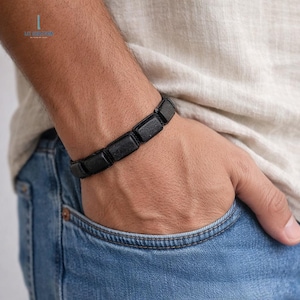 May include: A black rectangular stone bracelet is worn on a wrist. The bracelet is made of several dark, rectangular stones strung together. The person is wearing a light-colored shirt and blue jeans. The bracelet is a stylish accessory.