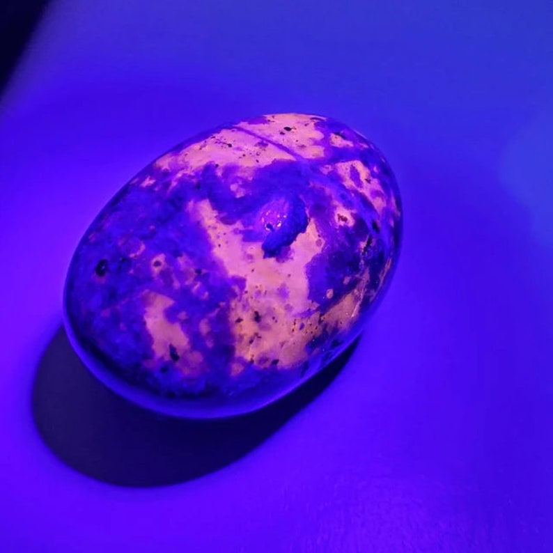 May include: A smooth, oval-shaped stone with a pattern of purple and cream hues. The stone is lit with a purple light, giving it a glowing appearance. The surface of the stone has a polished texture.