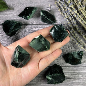 Puede incluir: Una colecci&oacute;n de piedras de obsidiana verde oscuro, con bordes &aacute;speros e irregulares. Las piedras se sostienen en una mano, mostrando su textura natural y v&iacute;trea. Ramas de lavanda seca en el fondo, agregando un elemento natural a la composici&oacute;n.