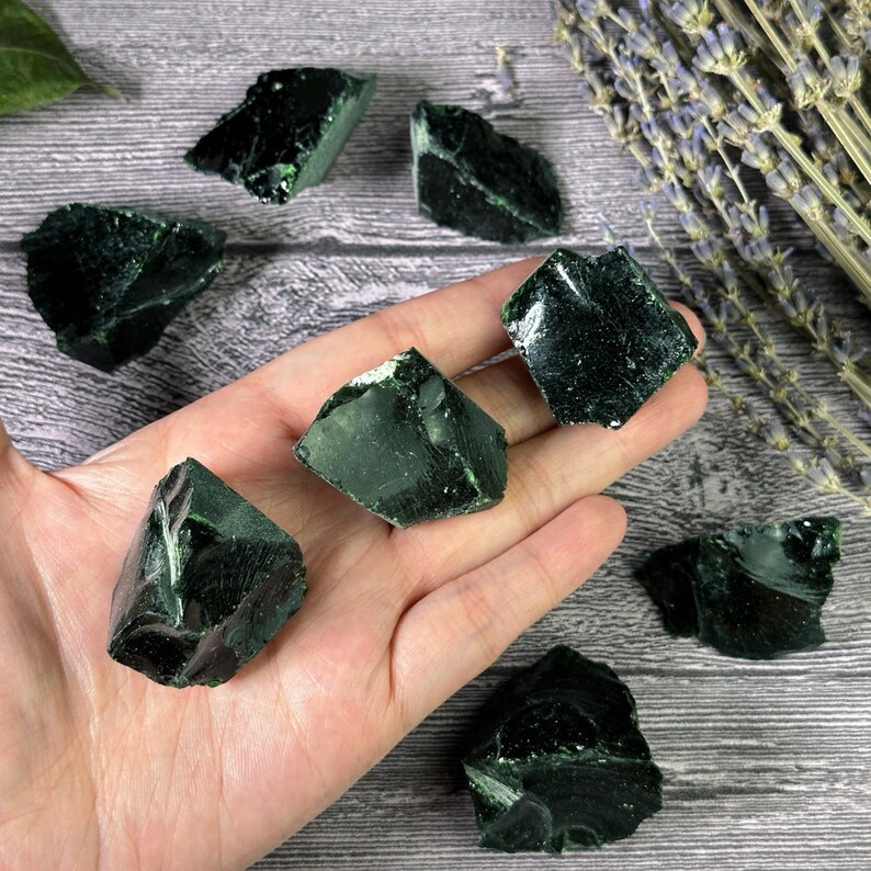 Puede incluir: Una colecci&oacute;n de piedras de obsidiana verde oscuro, con bordes &aacute;speros e irregulares. Las piedras se sostienen en una mano, mostrando su textura natural y v&iacute;trea. Ramas de lavanda seca en el fondo, agregando un elemento natural a la composici&oacute;n.