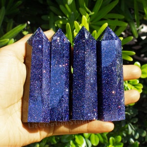 May include: Four blue sandstone obelisks with sparkling inclusions are held in a hand. The obelisks have a rectangular base and a pointed top. The background is a blurred view of green foliage.