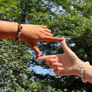 Pode incluir: Duas pulseiras de contas, uma com contas pretas, douradas e castanhas, a outra com contas rosa e douradas, s&atilde;o apresentadas nos pulsos. As m&atilde;os formam uma moldura contra um fundo de folhagem verde.