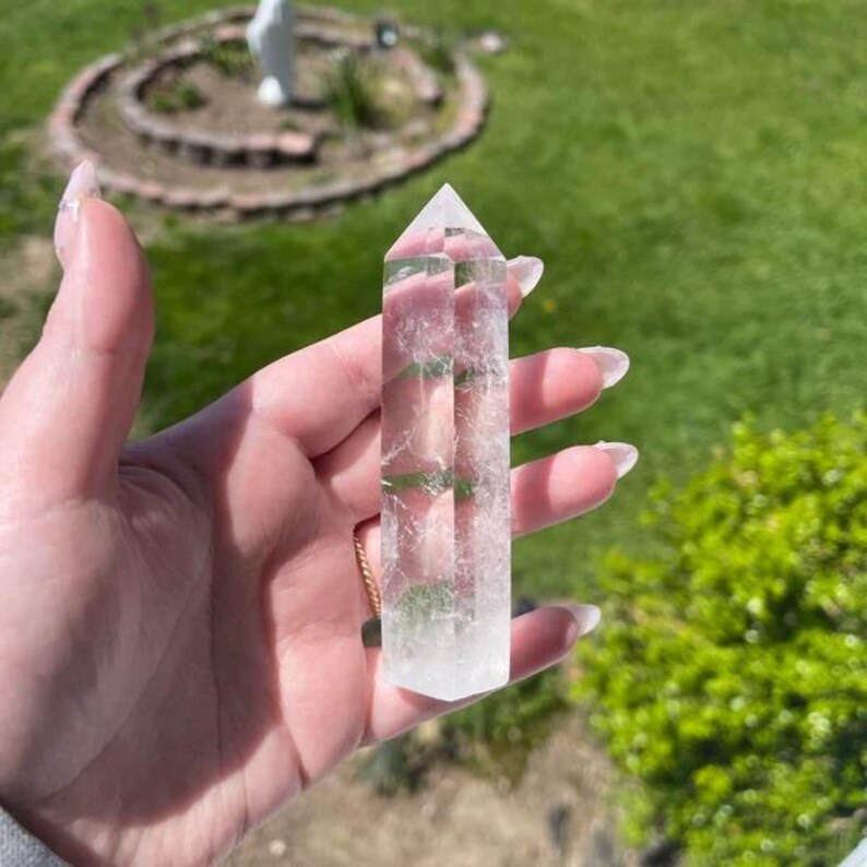 Puede incluir: Una punta de cristal de cuarzo transparente con doble terminaci&oacute;n. El cristal es liso y pulido, con una ligera apariencia nublada. El cristal se sostiene en una mano contra un fondo de hierba verde.