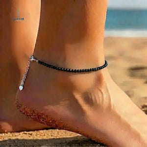 May include: A black beaded anklet with a silver-toned chain and heart charm. The anklet is worn on a foot on a sandy beach, with the ocean visible in the background. The anklet is delicate and simple.