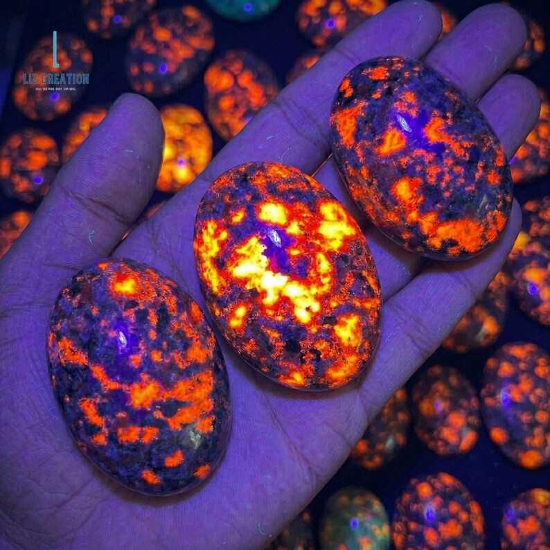 Puede incluir: Tres piedras ovaladas con un vibrante patr&oacute;n naranja y amarillo, sostenidas en una mano. Las piedras tienen una apariencia moteada con acentos azul oscuro y p&uacute;rpura. El fondo presenta m&aacute;s piedras iguales, creando una colecci&oacute;n de piedras &uacute;nicas.