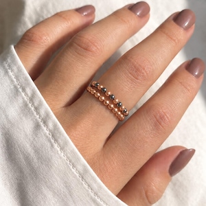 May include: Two stacked rings featuring small, round beads. One ring consists of metallic rose gold-colored beads, while the other ring is composed of alternating dark grey and rose gold-colored beads, creating a layered, beaded appearance.