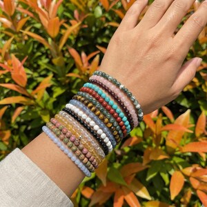 以下が含まれることがあります： ピンク、青、緑、茶色など、さまざまな色のビーズブレスレットのコレクション。 ブレスレットは手首に重ねられ、さまざまな宝石ビーズが展示されています。 背景には緑とオレンジの葉があります。
