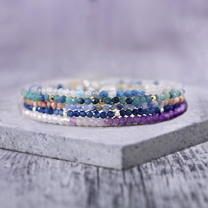 May include: A stack of colorful gemstone bracelets in shades of blue, purple, and pink. The bracelets are made of small, round beads and are layered on top of each other.
