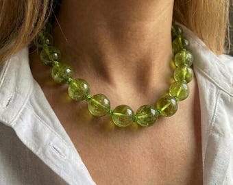 May include: A beaded necklace featuring large, round, translucent green beads. The beads are strung together with a matching green cord. The necklace is worn against a white shirt.