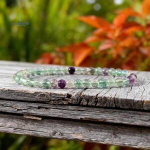 May include: A beaded bracelet featuring translucent green, purple, and clear fluorite stones. The bracelet is strung on an elastic band and rests on a weathered wooden surface. The background shows blurred green and orange foliage.