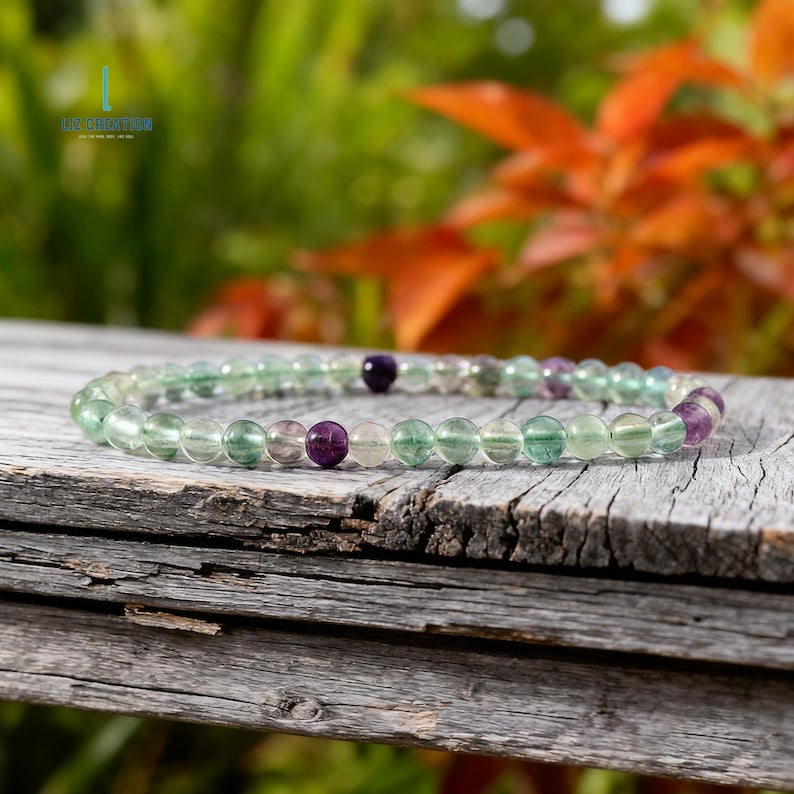 May include: A beaded bracelet featuring translucent green, purple, and clear fluorite stones. The bracelet is strung on an elastic band and rests on a weathered wooden surface. The background shows blurred green and orange foliage.