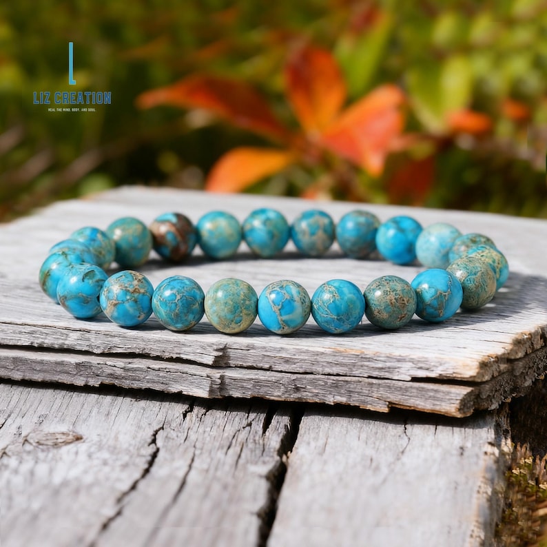 Pu&ograve; includere: Un braccialetto di perline turchesi con un mix di tonalit&agrave; blu e marroni. Le perline rotonde sono infilate insieme, appoggiate su una superficie di legno stagionato. L'immagine include il testo "LIZ CREATION".