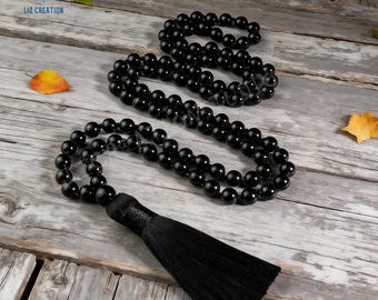 May include: Three strands of black beaded necklaces with a black tassel. The beads are round and shiny. The necklaces are displayed on a weathered wooden surface with a few leaves.