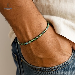 May include: A beaded bracelet featuring small, cylindrical turquoise-colored stones with natural veining, interspersed with thin, dark metallic spacers. The bracelet is worn around the wrist, showcasing a repeating pattern of blue-green beads.