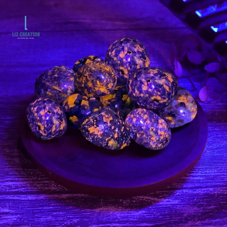 May include: A selection of polished stones with a speckled pattern in purple, black, and orange, presented on a dark purple plate. The stones have a distinctive, textured look. The image is lit with purple light, and the text "LIZ CREATION" is visible.