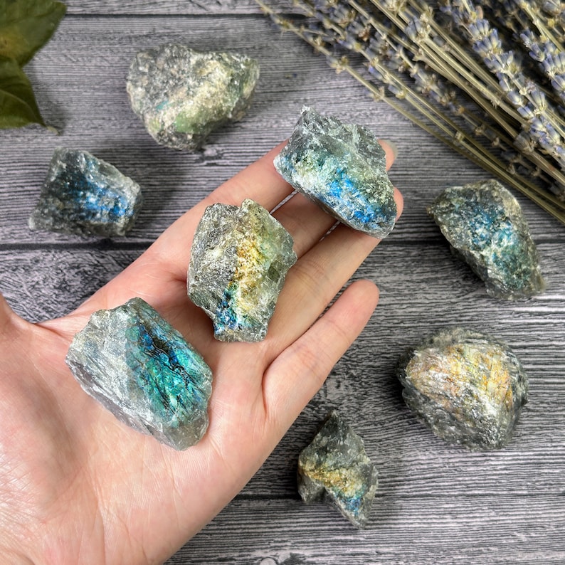 May include: A collection of raw labradorite crystals, showcasing iridescent flashes of blue, green, and gold. The rough-cut stones are held in a hand, with dried lavender sprigs in the background. The crystals vary in size and shape, with a natural, earthy aesthetic.