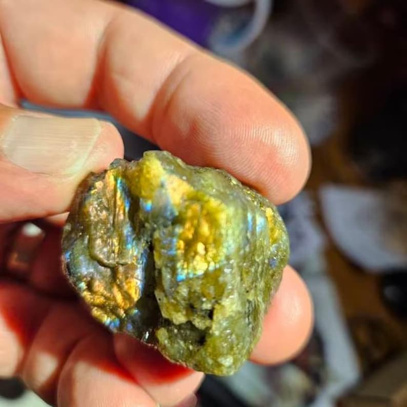 May include: A rough-cut labradorite gemstone with a mix of green, gold, and blue iridescence. The stone has a textured surface and is held in a hand, showcasing its natural, raw form. The colors shift with the light.