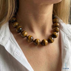 May include: A necklace featuring large, round tiger's eye beads. The beads are a warm brown color with dark brown stripes, strung together on a dark cord. The necklace is worn against a white shirt.