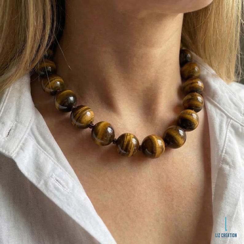May include: A necklace featuring large, round tiger's eye beads. The beads are a warm brown color with dark brown stripes, strung together on a dark cord. The necklace is worn against a white shirt.