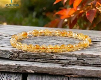 May include: A citrine beaded bracelet rests on a weathered wooden surface. The bracelet features round, translucent, golden-yellow beads. The background includes blurred greenery and reddish-orange foliage. The text "LIZ CREATION" is visible in the upper left corner.