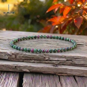 Op de afbeelding: Een kralenarmband bestaande uit kleine, ronde edelstenen in groen en rood. De armband ligt op een verweerd houten oppervlak, met een vage natuurlijke achtergrond. Het sieraad is een armband.