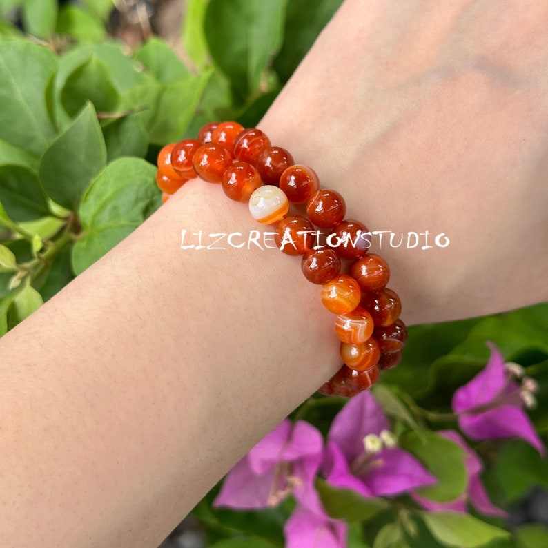 Puede incluir: Dos pulseras de cuentas con piedras de &aacute;gata naranja y blanca. Las pulseras se llevan en la mu&ntilde;eca de una persona. Las cuentas son redondas y tienen un acabado pulido. Las pulseras se colocan sobre un fondo de hojas verdes y flores moradas. El texto "lizcreationsstudio" es visible.