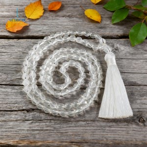 Op de afbeelding: Een heldere kwarts kristallen mala kralenketting met een witte kwast, op een verweerd houten oppervlak. De ronde, doorschijnende kralen vormen de ketting. Een spiritueel of meditatie accessoire.