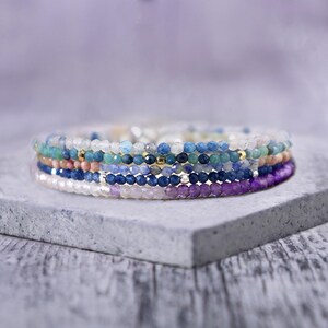May include: A stack of five delicate beaded bracelets featuring faceted gemstones in shades of light blue, teal, peach, dark blue, and purple. The bracelets are accented with small metallic gold and silver beads interspersed throughout the strands.