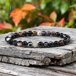 May include: A bracelet made of round, polished gemstones in black, brown, and cream. The bracelet is displayed on a weathered wooden surface, with a blurred background of green and orange leaves. The bracelet is a piece of jewellery.