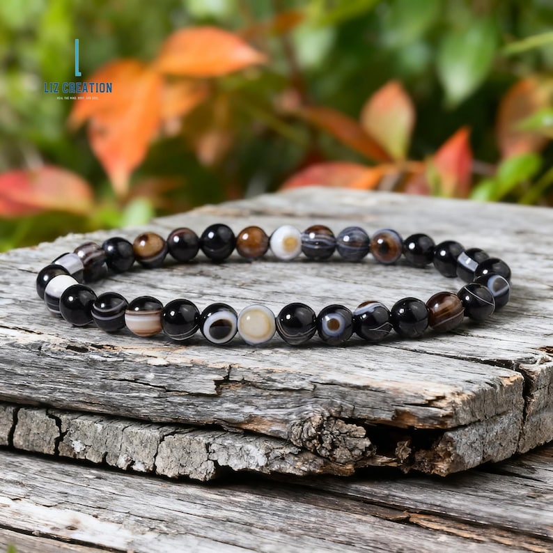 May include: A bracelet made of round, polished gemstones in black, brown, and cream. The bracelet is displayed on a weathered wooden surface, with a blurred background of green and orange leaves. The bracelet is a piece of jewellery.