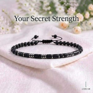 May include: A black beaded bracelet featuring a series of matte black cylindrical beads and small metallic silver spacers arranged in a pattern, with a sliding knot closure. The text "Your Secret Strength" is printed above the bracelet.
