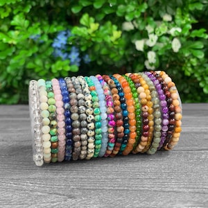 May include: A collection of beaded bracelets in various colors, including clear, pink, blue, green, and brown. The bracelets are made of small, round beads and are stacked together. The background is a blurred mix of green foliage.