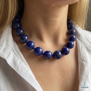 May include: A necklace featuring large, spherical lapis lazuli beads in a deep blue color with natural gold-colored flecks. The beads are strung together in a single strand, creating a bold statement piece that rests around the neck area.
