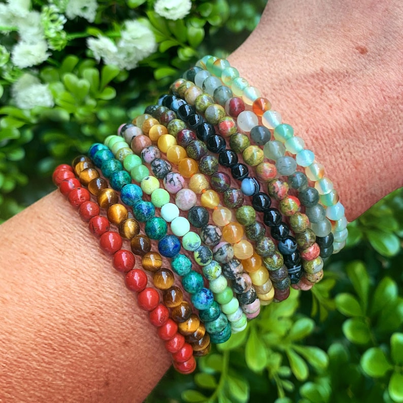 Op de afbeelding: Een stapel van 14 kleurrijke kralenarmbanden om de pols van een persoon. De armbanden zijn gemaakt van verschillende edelstenen, waaronder rood, oranje, geel, groen, blauw en paars.