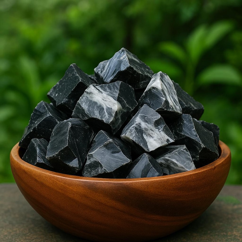 Puede incluir: Un cuenco de madera lleno de piedras de obsidiana negra. Las piedras tienen una forma rugosa y angular con vetas blancas. El cuenco es de madera pulida, con un fondo verde borroso.