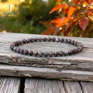 May include: A circular beaded bracelet composed of small, round, dark brown stones featuring white, snowflake-like speckled patterns throughout each bead. The bracelet is arranged in a continuous loop, showcasing a uniform size and texture for every individual stone bead.