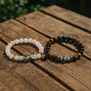 Pode incluir: Duas pulseiras de contas sobre uma superf&iacute;cie de madeira. Uma tem contas brancas e rosa com detalhes dourados e pingentes de lua. A outra tem contas marrons, pretas e prateadas. Estas pulseiras s&atilde;o uma joia.