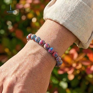 May include: A beaded bracelet featuring round, multi-colored beads in shades of blue, red, and tan. The bracelet is worn on a wrist, with the brand name "LIZ CREATION" visible in the background.