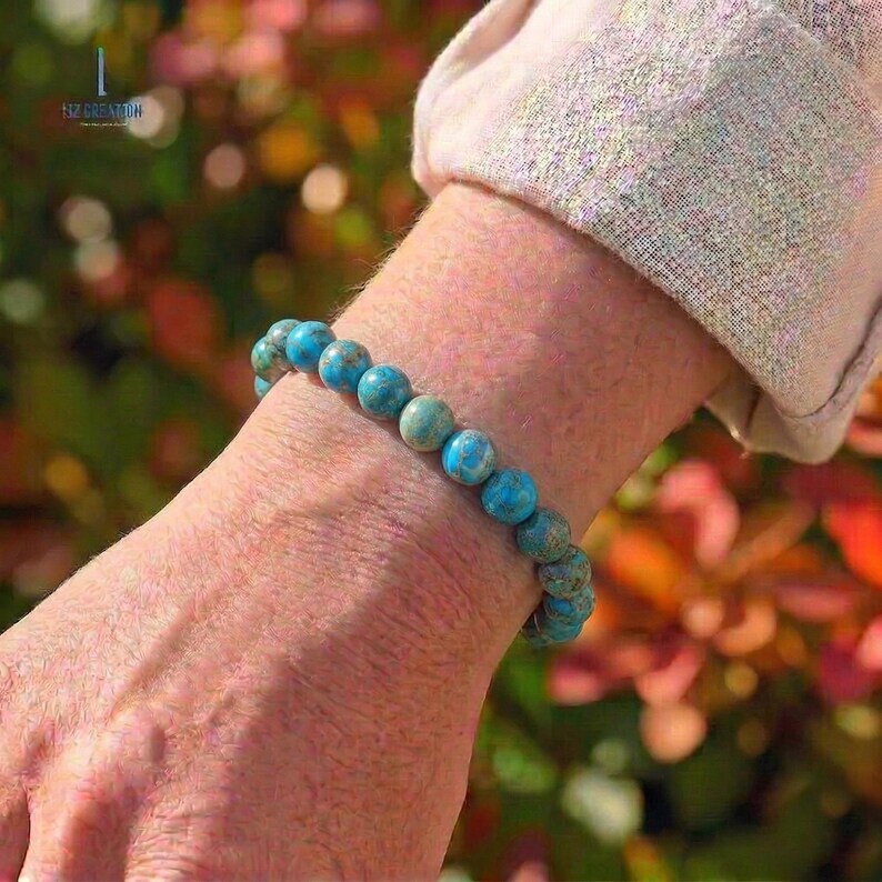 Pu&ograve; includere: Un braccialetto di perline turchesi. Il braccialetto &egrave; composto da perline rotonde e lucide nei toni del turchese e del verde acqua. Il braccialetto &egrave; indossato su un polso, con uno sfondo sfocato di fogliame verde e rosso.