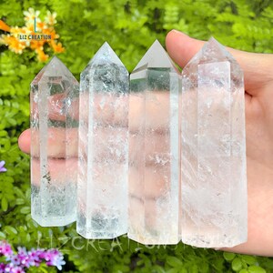 Puede incluir: Cuatro puntas de cristal de cuarzo transparente, tambi&eacute;n conocidas como varitas, est&aacute;n dispuestas en fila. Los cristales son lisos y transl&uacute;cidos, con un ligero brillo. Los cristales se sostienen en una mano, con un fondo verde de follaje.