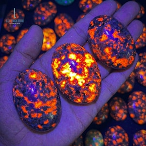 May include: Three oval-shaped sodalite stones with vibrant orange and yellow patterns, held in a hand. The stones have a speckled appearance with dark blue and purple hues. The background features more of the same stones.