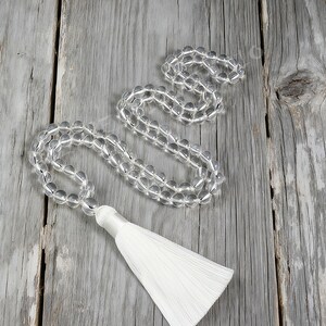 Op de afbeelding: Transparante kwarts kristallen kralenketting met een witte kwast. De ronde kralen zijn transparant. De kwast is gemaakt van witte draden. De ketting wordt gepresenteerd op een verweerd houten oppervlak. Ongeveer 50 cm lang.