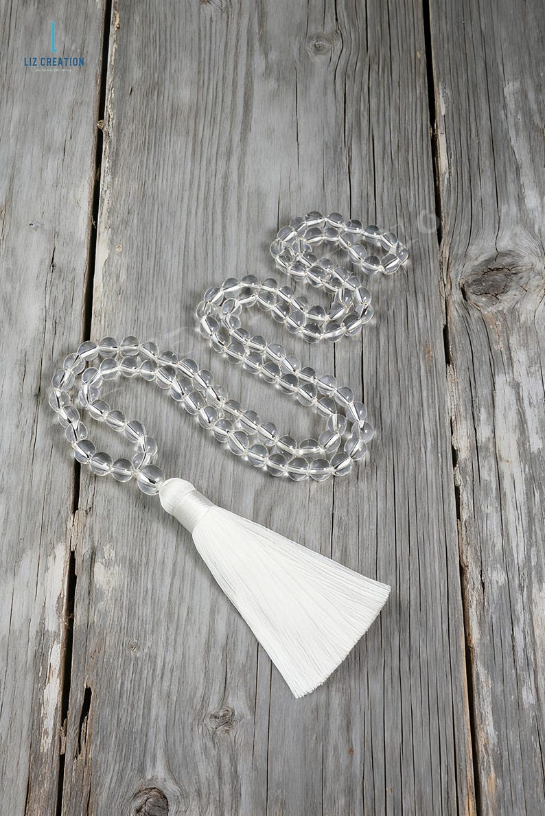 Op de afbeelding: Transparante kwarts kristallen kralenketting met een witte kwast. De ronde kralen zijn transparant. De kwast is gemaakt van witte draden. De ketting wordt gepresenteerd op een verweerd houten oppervlak. Ongeveer 50 cm lang.