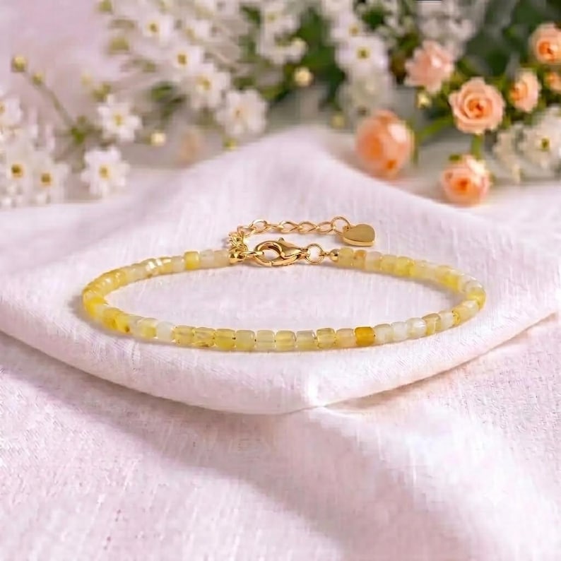 May include: A delicate bracelet featuring small, translucent yellow and white beads strung together. The piece includes a gold-toned metal lobster clasp, an adjustable chain extension, and a tiny heart-shaped charm attached to the end of the chain.