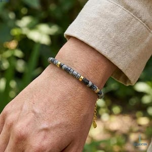 Puede incluir: Una pulsera de cuentas con cuentas de piedra grises y negras intercaladas con espaciadores dorados. La pulsera está en una cadena dorada con un pequeño dije en forma de corazón. La pulsera se usa en una muñeca.