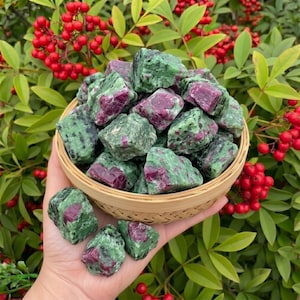 May include: A hand holds a woven basket filled with rough-cut zoisite stones with ruby inclusions. The stones are a mottled green with deep red patches. The background features green foliage and red berries.